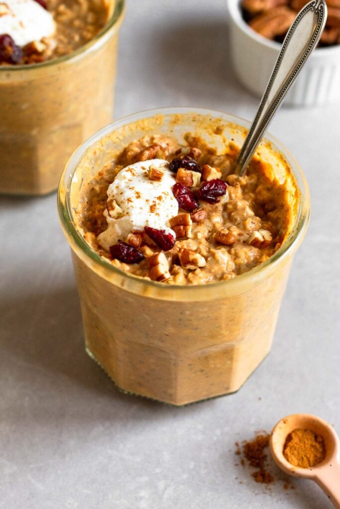 Pumpkin overnight oats in a jar topped with greek yogurt, chopped pecans, dried cranberries, and cinnamon. A spoon is also in the jar. Around the jar is a ramekin of pecans and another jar of pumpkin overnight oats.