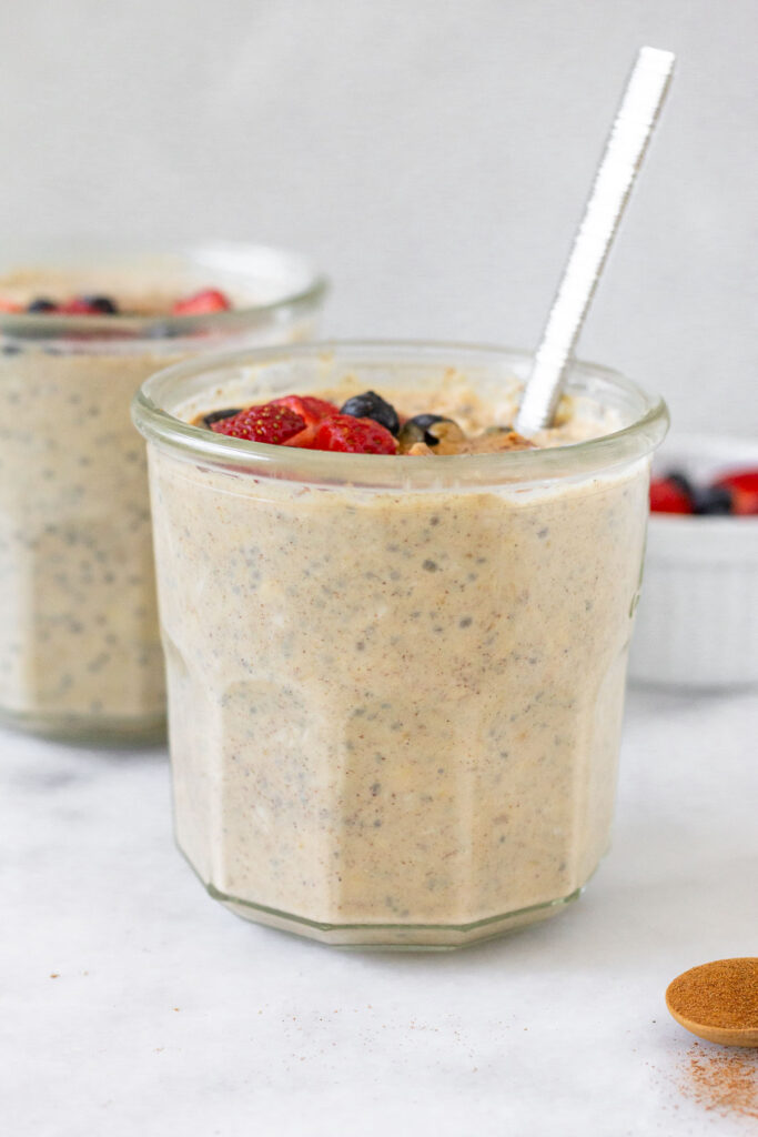 Two jars of overnight oats with protein powder, with one being behind the other. They are topped with berries and a spoon is in the jar in the front. In front of the jar is a teaspoon of cinnamon and off to the back is a dish of berries.